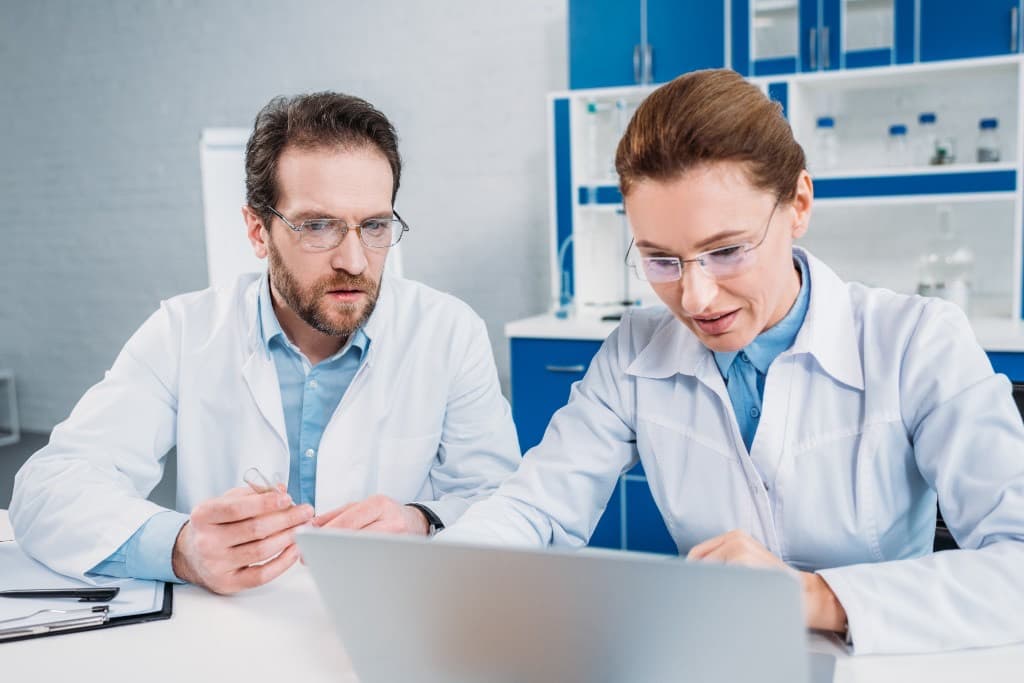 Two scientists reviewing results on a laptop, one holding a sample vial